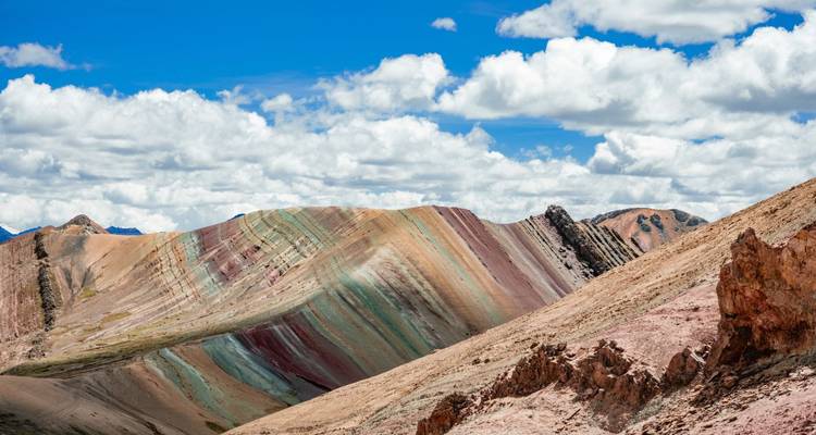 Colour-banded slopes of Rainbow Mountain curve beneath a bright sky filled with cumulus clouds.