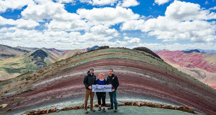 Three travellers hold a banner on a ridge of Rainbow Mountain with multicoloured ranges behind.