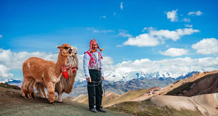 Local man in traditional clothes stands with a decorated alpaca against snow-capped Andes peaks.