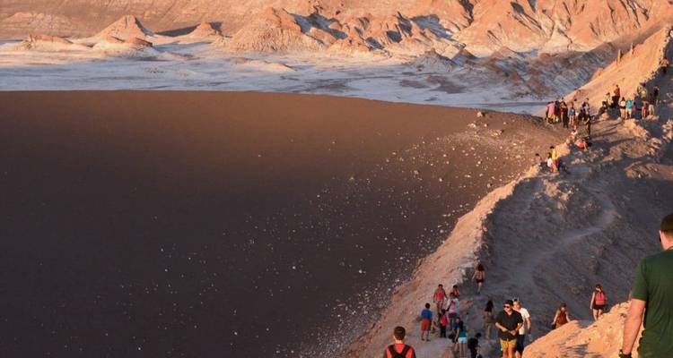 Hikers trek along a high ridge of red sand overlooking the vast desert dunes of Valle de la Luna at dusk.