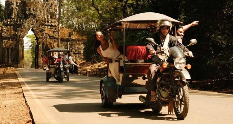 Tuk-tuk convoy drives through an ancient stone gate adorned with large carved faces in Cambodia.