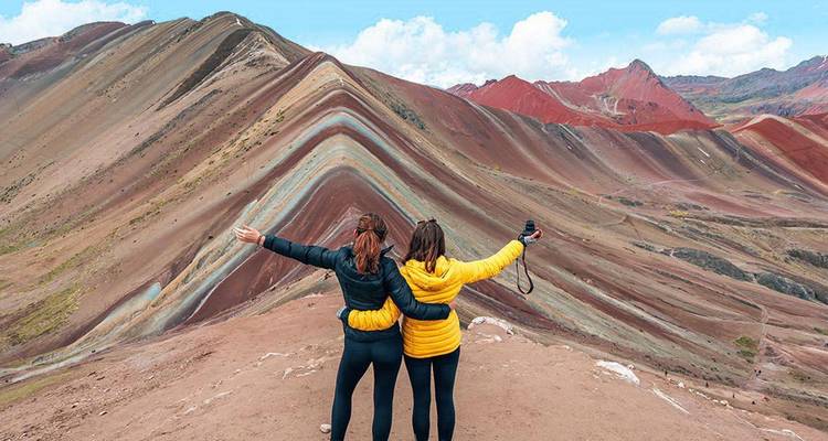 Two hikers with outstretched arms admire the vibrant striped slopes of Peru’s Rainbow Mountain.