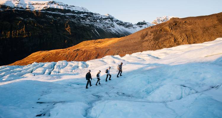 Vier wandelaars steken een helderblauwe gletsjer over met besneeuwde toppen en gouden heuvels op de achtergrond
