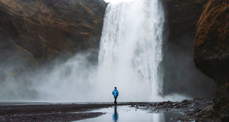 Eenzame reiziger staat vol ontzag voor het bulderende gordijn van waterval Skógafoss gehuld in mist
