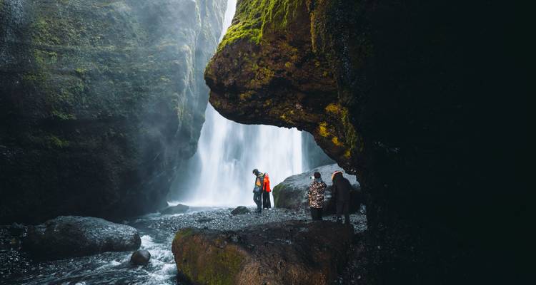 Reizigers staan op natte rotsen in een met mos bedekte kloof en kijken naar een krachtige waterval die tussen hoge kliffen naar beneden stort
