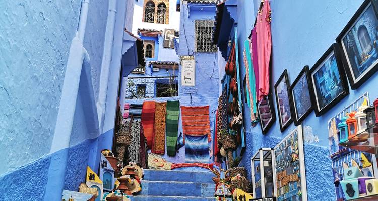 Escalier bleu escarpé à Chefchaouen bordé de tapis colorés, d'artisanat et de photos encadrées à vendre.