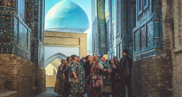 Grupo de mujeres uzbekas vestidas tradicionalmente escuchan a la guía en un pasadizo de azulejos azules.
