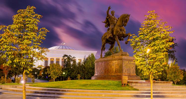 Estatua ecuestre de bronce de Amir Timur se alza frente a un edificio gubernamental iluminado bajo un crepúsculo púrpura intenso.