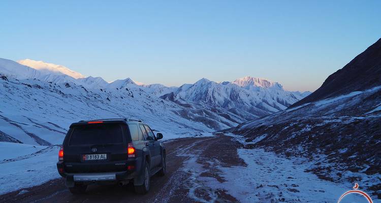 Een SUV rijdt over een sneeuwige onverharde bergpas met rondom zich hoog oprijzende, wit besneeuwde bergen in het zachte licht van de dageraad.