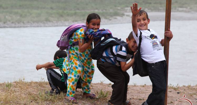 Lachende kinderen met rugzakken zwaaien speels naast elkaar aan de oever van een rivier op een stoffig pad.