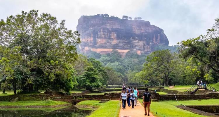 Visitors walk along water gardens toward the massive Sigiriya rock fortress rising in mist.