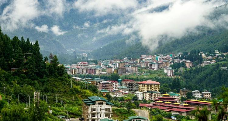 Misty mountain valley filled with clustered Bhutanese buildings surrounded by lush green forests.