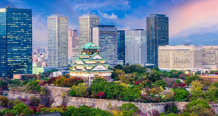 El Castillo de Osaka se alza entre rascacielos modernos y árboles otoñales bajo un cielo vespertino lleno de colores.