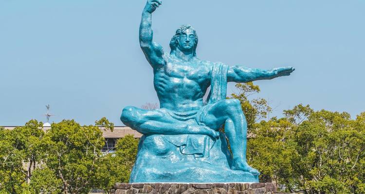 La estatua azul de la Paz de Nagasaki se encuentra contra un cielo despejado sobre exuberante verdura.