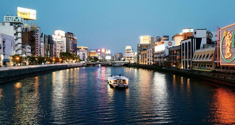 Un barco solitario se desliza por un río iluminado por luces de neón rodeado de edificios modernos en Fukuoka al atardecer.