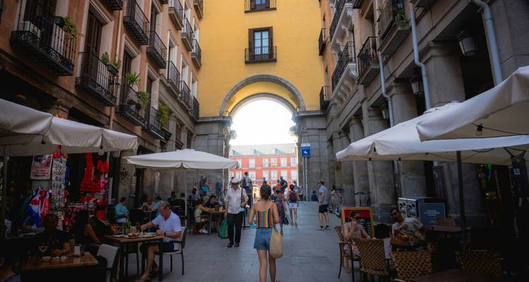 Callejón cubierto y bullicioso se abre a la Plaza Mayor de Madrid; los comensales se sientan bajo sombrillas mientras los turistas exploran.
