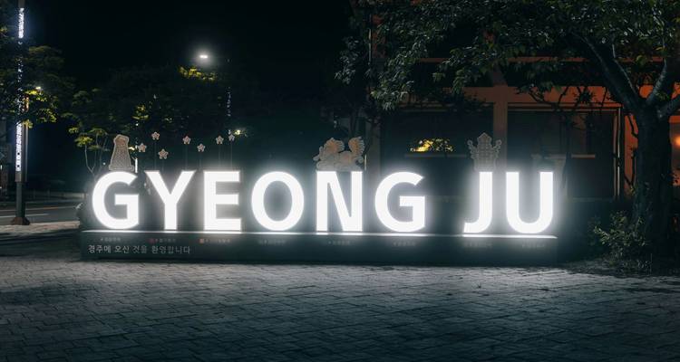 Cartel iluminado de bienvenida "GYEONG JU" brillando de noche en una plaza de la ciudad.
