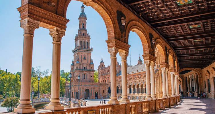 Gewelfde galerij die de indrukwekkende toren van Plaza de España in Sevilla omlijst, met ochtendzon en groen park erachter.
