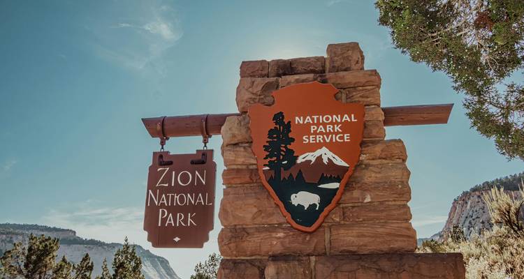 Letrero de entrada de piedra y madera del Parque Nacional Zion contra un cielo despejado.