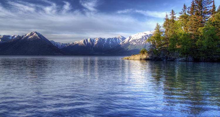 Still alpine lake with evergreen peninsula reflects distant snow-dusted mountain range beneath dramatic sky.