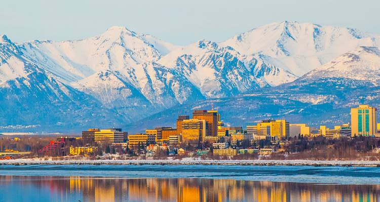 Anchorage skyline backed by snow-covered Chugach Mountains reflected in coastal waters at dusk.