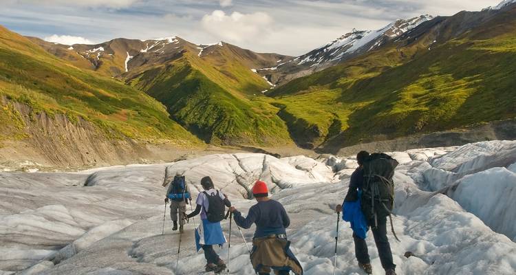 Adventurers trek across crevassed glacier with crampons toward green alpine valley in Alaska.