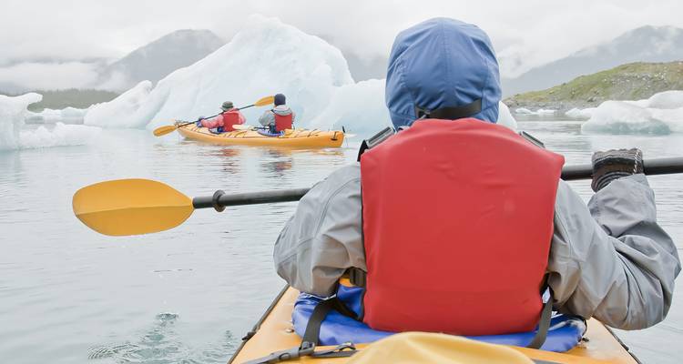 Kayakers paddle among blue icebergs in misty glacial lagoon viewed from another kayak perspective.