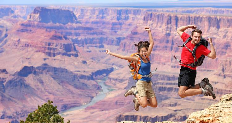 Jong stel met rugzakken springt blijde omhoog aan de rand van de Grand Canyon met de rivier zichtbaar ver beneden