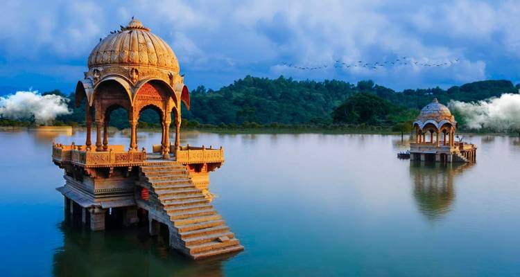 Ornate sandstone cenotaphs rising from tranquil lake waters with forested hills beyond near Jaisalmer.