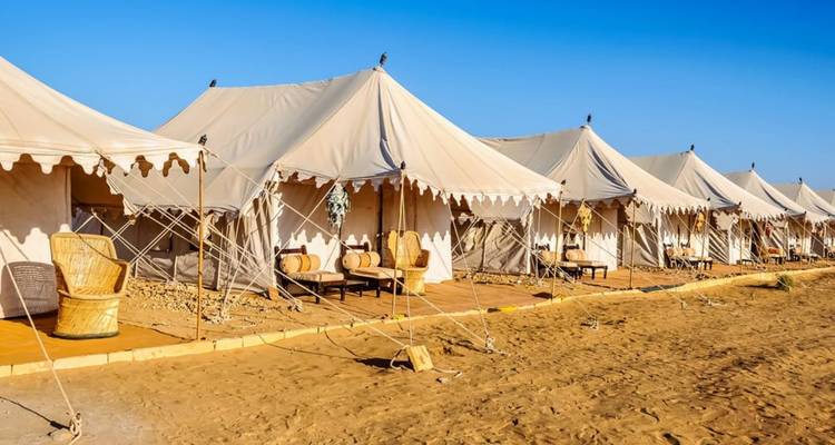 Row of luxury canvas tents with wicker chairs set on sandy desert ground under clear blue sky.