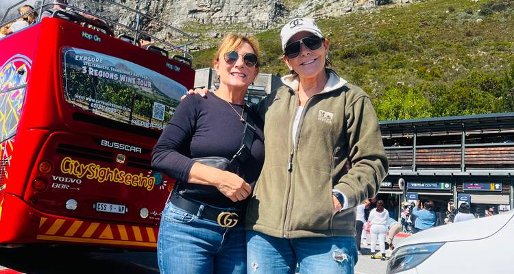 Two women posing in front of red open-top sightseeing bus at foot of rocky Table Mountain