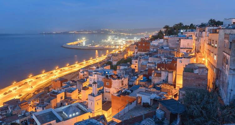 Evening cityscape of a coastal Moroccan town overlooking a lit-up harbor road.