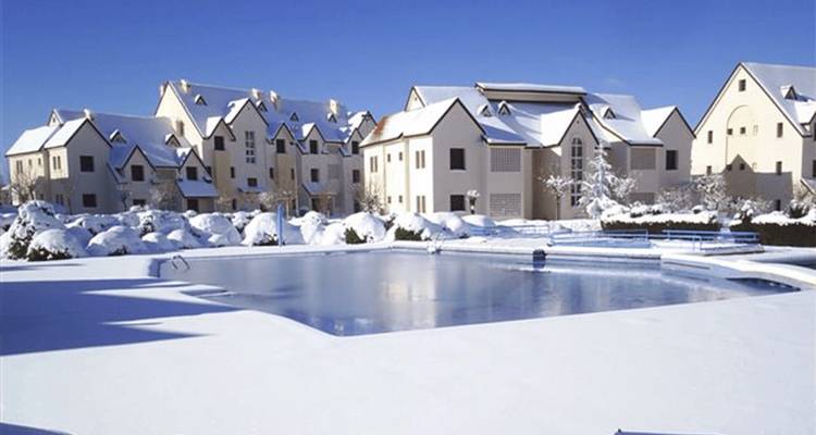 Snow-covered resort houses surrounding an outdoor pool under clear blue skies.