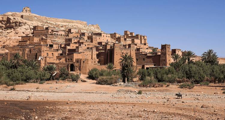 Ancient earthen fortress village rising above a dry riverbed with palm trees.