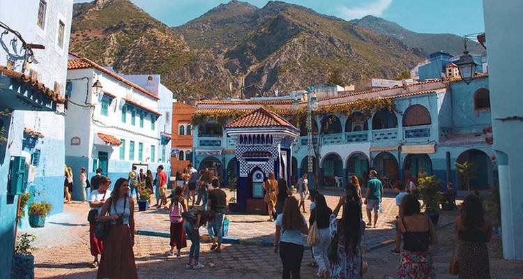 Busy blue-and-white medina square of Chefchaouen filled with tourists and overlooked by rugged mountains.