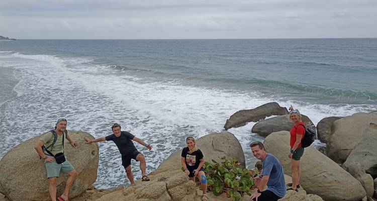 Les voyageurs posent sur des rochers en bord de mer avec des vagues qui s'écrasent derrière eux sous un ciel nuageux.