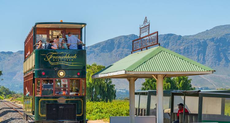 Historic green double-decker wine tram carrying passengers past vineyards with mountain backdrop.