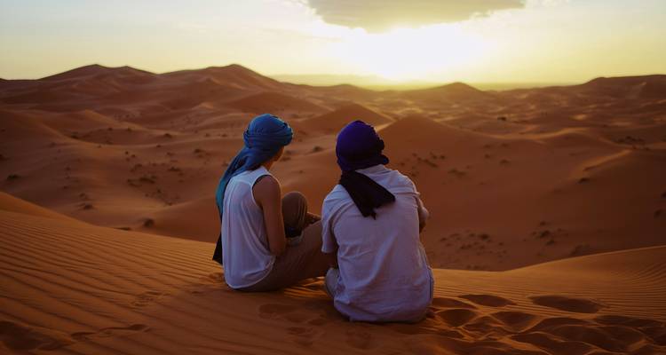 Deux voyageurs enveloppés de turbans sont assis sur une haute dune, regardant le soleil se coucher sur d'infinies vagues de sable.