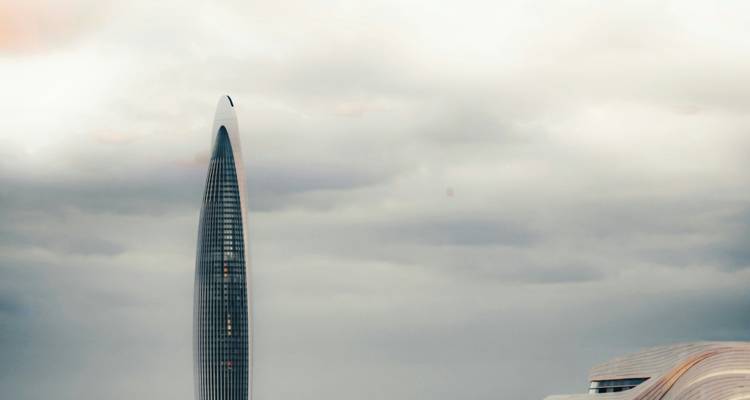 Un gratte-ciel élancé et imposant perce un ciel nuageux au-dessus de structures modernes basses.