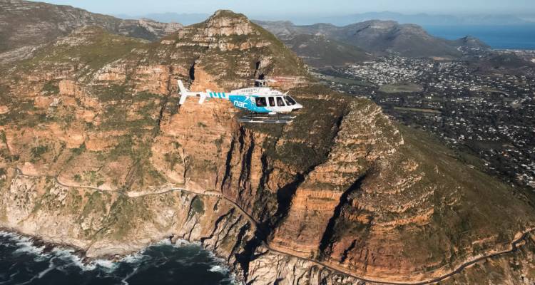 Blue and white helicopter flies past rugged sea cliffs and winding coastal road.