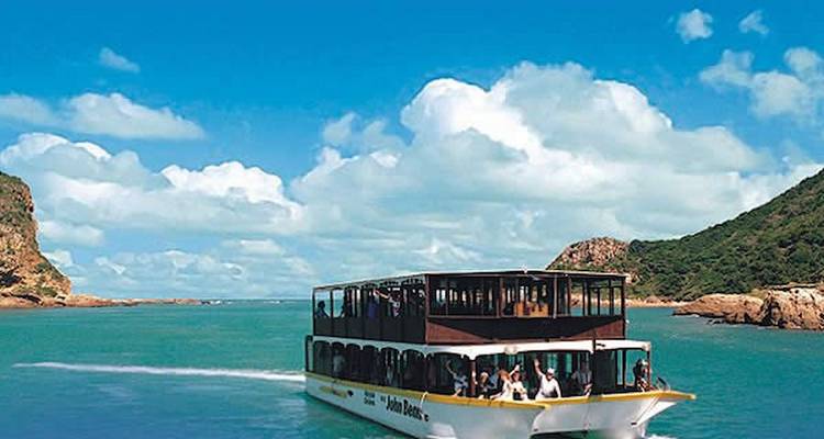 Two-level sightseeing boat cruises through emerald lagoon surrounded by green hills under blue sky.