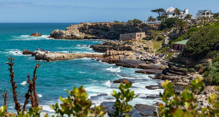 Rocky shoreline with crashing waves and cliff-top homes near vibrant turquoise sea.