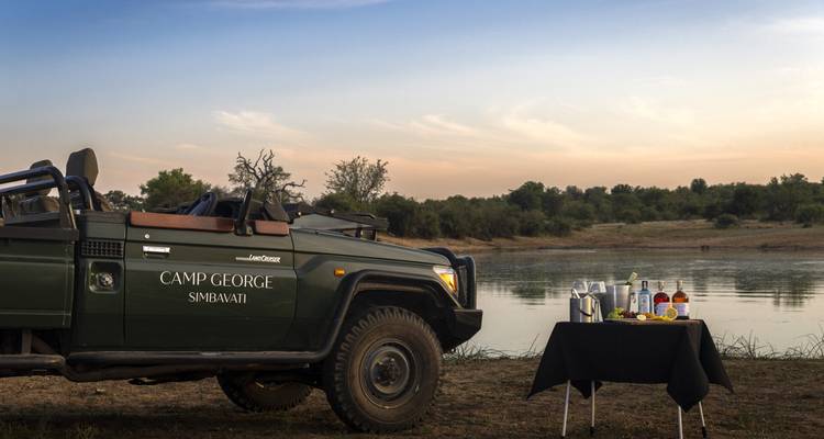 Open safari vehicle parked beside tranquil waterhole set for sundowner drinks in bushveld.
