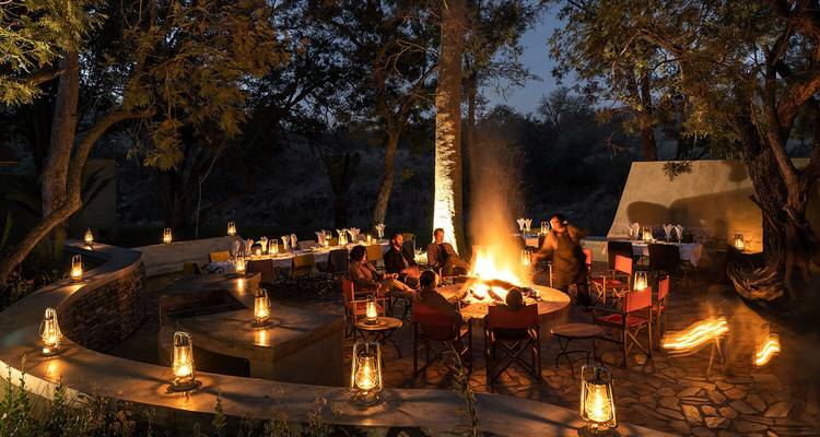Guests dine around campfire beneath lantern-lit trees creating warm ambience at night.