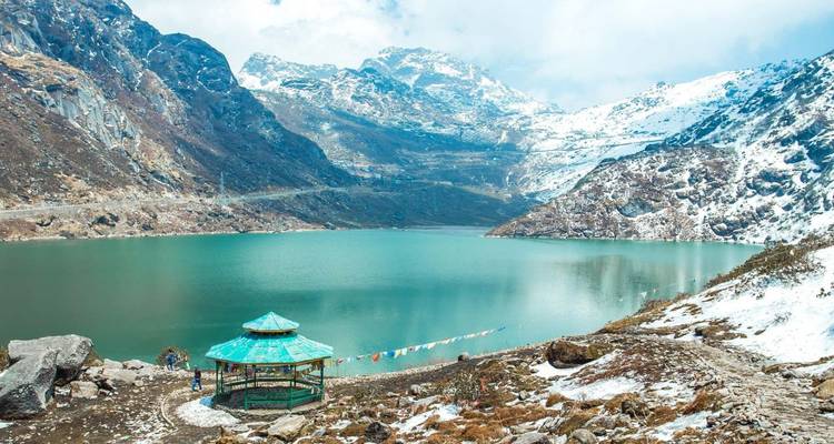 Turquoise alpine lake surrounded by snow-dusted peaks with a small green pavilion in the foreground.