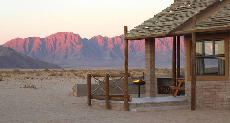 Rustic desert lodge with a small fire pit facing pink-hued mountain ranges at sunset in the Namib.