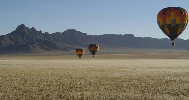Three colorful hot air balloons float above a vast grassy plain with rugged mountain silhouettes in Namibia’s clear morning light.