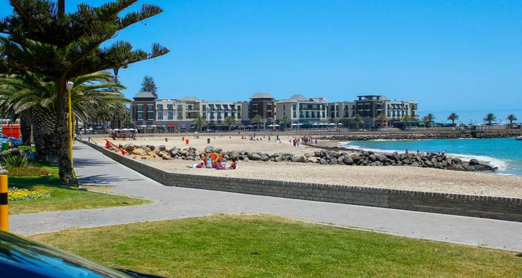 Sandy urban beach with visitors, palm trees and modern beachfront buildings under bright blue sky.
