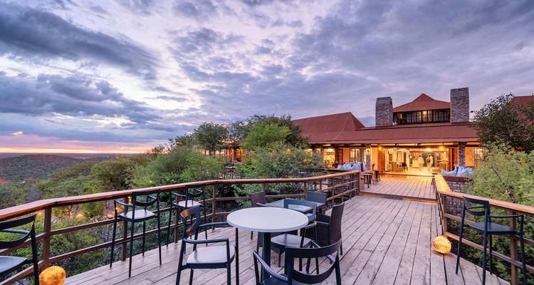 Wooden terrace with tables overlooking a lush valley and stylish lodge lit by warm evening light.