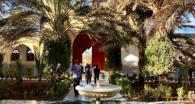 Los visitantes caminan a través de un patio exuberante con palmeras y una fuente de mármol hacia una puerta con arco rojo.